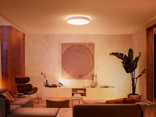 Ceiling-mounted round smart light fixture emitting warm illumination over a living room with seating, table, and decorative objects.