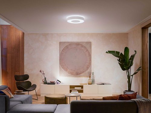 Ceiling-mounted circular smart light fixture emitting white illumination over a living room with furniture and decorative elements.
