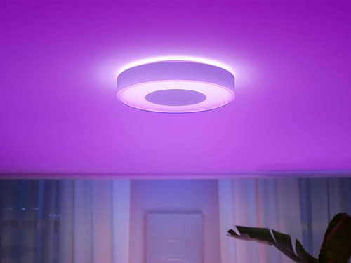 Round ceiling-mounted smart light emitting purple illumination onto the ceiling, positioned above a window with curtains.