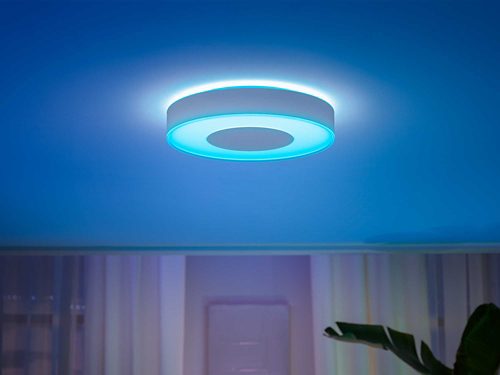 Round ceiling-mounted smart light fixture emitting blue illumination onto the ceiling in a modern room with curtains and a plant.