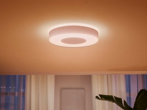 Round ceiling-mounted smart light fixture emitting diffused white light, installed in a room with curtains and a plant visible below.
