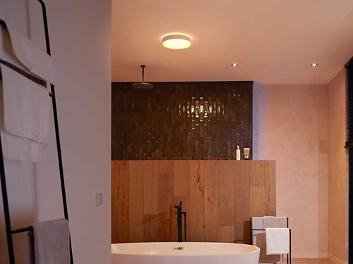 Ceiling-mounted round smart light fixture illuminated above bathtub, casting light onto wood panel and tiled wall in bathroom.