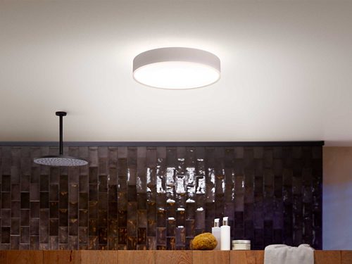 Round ceiling-mounted smart light fixture emitting bright illumination above a tiled wall and countertop with assorted bathroom items.