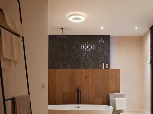 Circular ceiling light fixture mounted above a bathtub, emitting white light onto the bathroom with wood and tile surfaces.