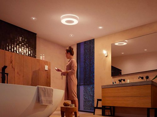 Person in a bathroom illuminated by ceiling and wall lights, standing near a bathtub and wooden vanity, holding an item in hand.