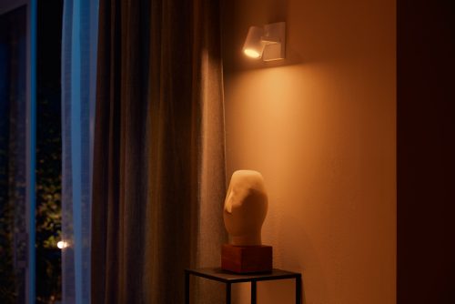 White single spot light fixture mounted on wall, illuminating a sculpture on a stand beside closed curtains.