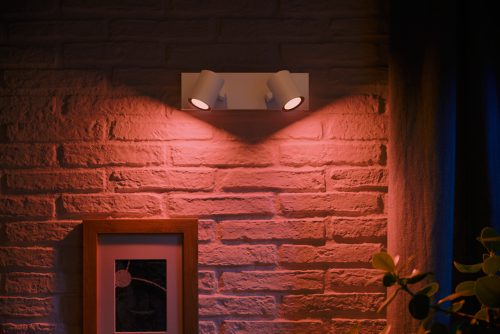 White plate spiral spotlight fixture mounted on brick wall, casting colored light onto wall above framed artwork and nearby curtain.