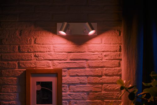 Wall-mounted spiral aluminium spotlight fixture projecting colored light onto a brick wall above a framed picture and curtain.