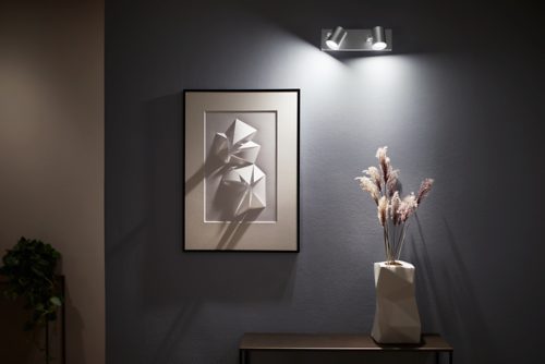 Aluminium spiral plate spotlight mounted on wall, illuminating framed geometric art and a vase with dried plants on a console table.