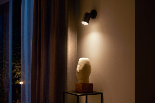 Black single spot light mounted on wall, illuminating a white sculpture placed on a stand beneath the fixture.