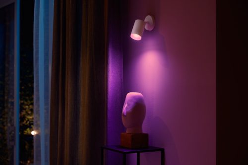 White single spot wall light mounted above a sculpture, casting purple illumination on the wall and object below.