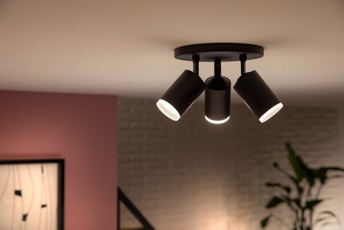 Black ceiling-mounted spotlight fixture with three adjustable heads, illuminating a room with a visible brick wall and plant in background.
