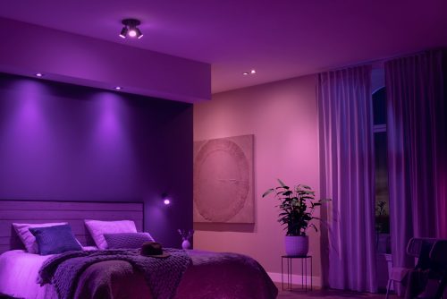 Bedroom illuminated by ceiling spotlights and wall lights with a purple glow, featuring a bed, plant, curtains, and textured artwork.