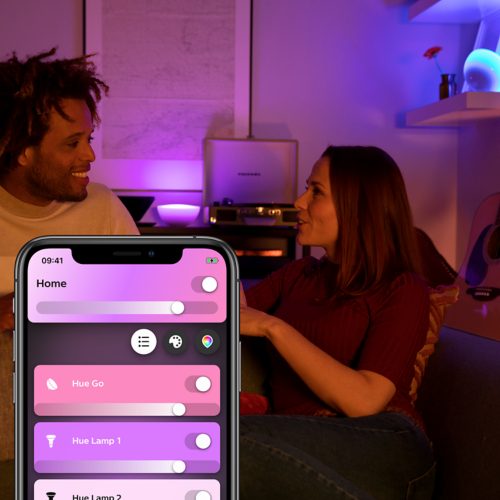 Two people sitting on a sofa adjust smart lighting via a mobile app, with purple and blue lights illuminating a living room.