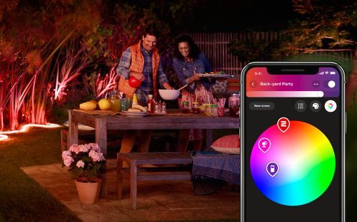 Group preparing food at a backyard table with colorful smart lighting and app interface showing light scene selection on a smartphone.