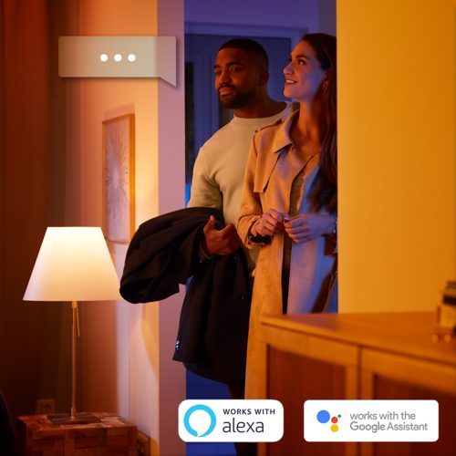 Two people entering a warmly lit living room, standing near a table lamp smart assistant logos are visible at the bottom.