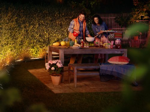 Two people preparing food at an outdoor table in a garden, supported by warm lighting along the hedge and seating area.