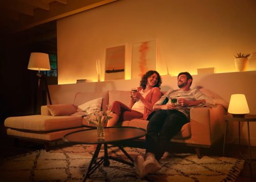 Two people sitting on a living room sofa, enjoying drinks with warm ambient smart lighting illuminating the space.
