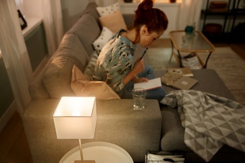 Person writing in a notebook on a living room sofa, illuminated by a table lamp with soft white light.