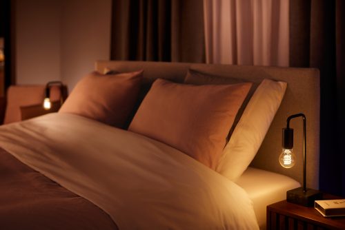 A filament-style smart bulb in a black table lamp placed on a bedside table, emitting warm white light onto the bed.