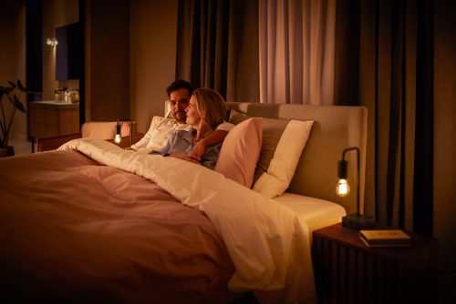 Two people relaxing in bed in a softly lit bedroom with warm dimmed bulbs on bedside tables enhancing the cozy setting.