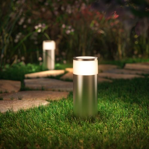 A cylindrical outdoor light with metallic finish installed in grass, illuminating nearby stone pathway in a garden setting.