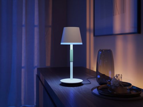 A white portable table lamp is placed on a dark wooden surface, emitting light while positioned on its charging base.