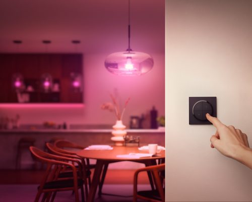 Finger pressing a black wall-mounted dial switch, likely to adjust smart lighting in a dining area with purple ambient light.