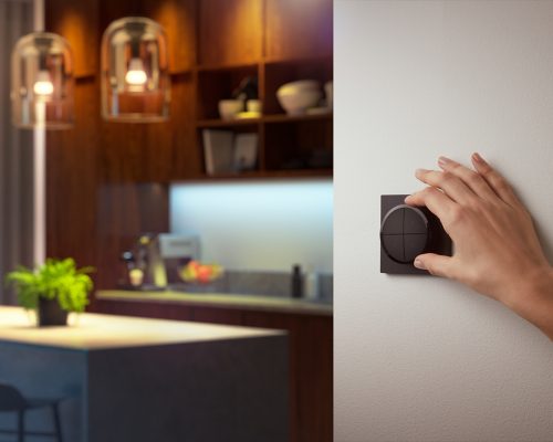 Hand rotating a black wall-mounted dial switch, likely for adjusting smart lighting settings, in a modern kitchen environment.