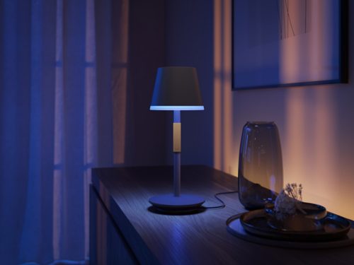 Black portable table lamp on charger placed on a wooden surface, emitting blue light onto nearby vase and decorative tray.