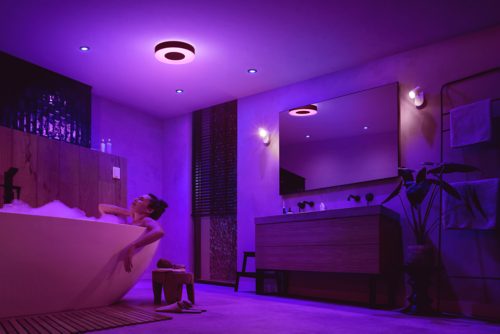Person relaxing in a bathtub in a modern bathroom, illuminated by purple ceiling and wall smart lights.