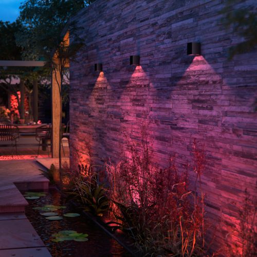 Black outdoor wall lights mounted on stone wall, casting colored downward beams onto plants and water feature below.