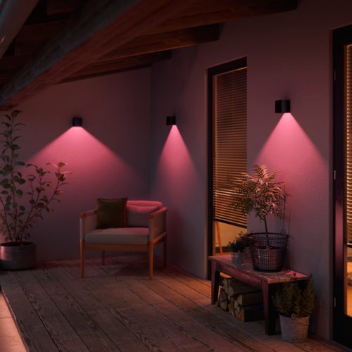 Outdoor wall-mounted black rectangular lights casting downward pink illumination onto a patio wall, highlighting nearby plants and furniture.