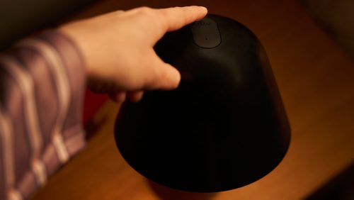 Finger pressing the top button on a black bedside table lamp, likely to operate or adjust the lamp's settings.