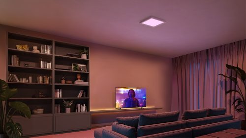 Square ceiling panel light emitting soft colored illumination above a living room setup with shelves, sofa, and television.