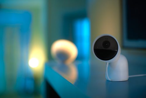 White wired security camera on a desktop stand placed on a cabinet, lens facing forward with cable visible on the surface.