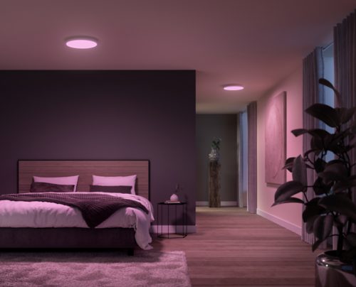 Round ceiling-mounted smart panel light emitting pink light, positioned above a bed in a modern bedroom with minimal decor.