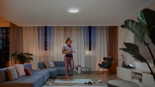 Person standing on a yoga mat in a living room, illuminated by a round ceiling light, with workout accessories nearby.