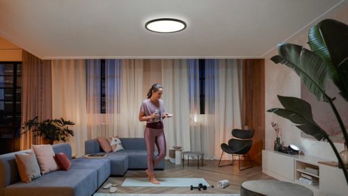 Person in workout attire stands on a yoga mat in a living room, illuminated by a round ceiling light above.