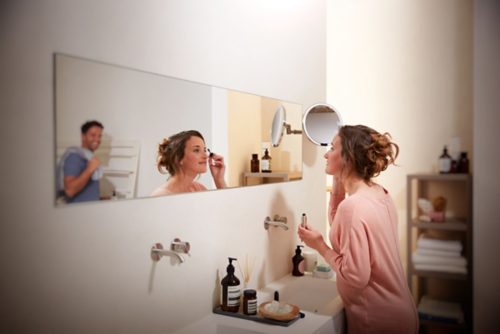 A person applying makeup at a bathroom sink, illuminated by soft lighting, with toiletries and towels visible in the background.