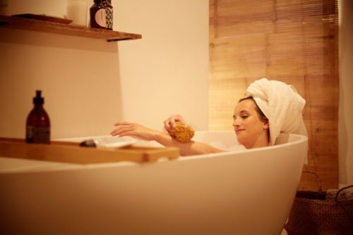 Person relaxing in a bathtub with a towel wrapped on their head, using a sponge, under soft warm lighting in a bathroom setting.