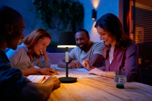 Group of people gathered outdoors around a table, using a portable black table lamp to read a map and plan together.