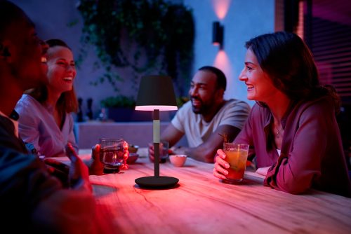 A group of people share drinks at an outdoor table, illuminated by a portable black table lamp with a pink light effect.