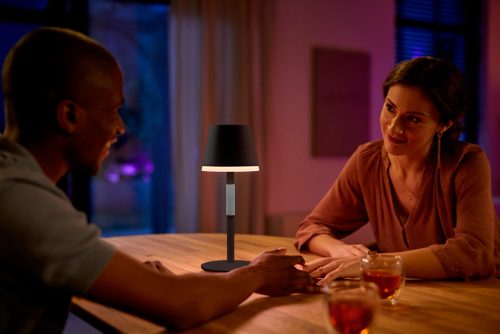 Two people seated at a table sharing a drink, illuminated by a black portable table lamp with warm light in a dining room setting.