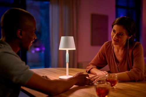 Two people share a meal at a wooden dining table, illuminated by a white portable table lamp in a softly lit indoor setting.