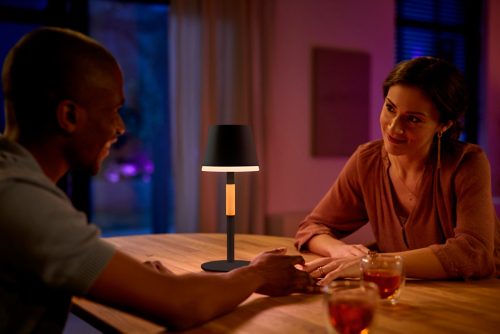 Two people sharing a meal at a wooden table, illuminated by a portable black table lamp with warm light in a cozy indoor setting.