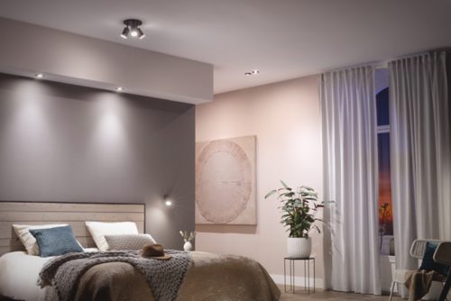 Ceiling-mounted black spotlight fixture illuminating a bedroom, with directed white light on walls and bed, enhancing visibility.