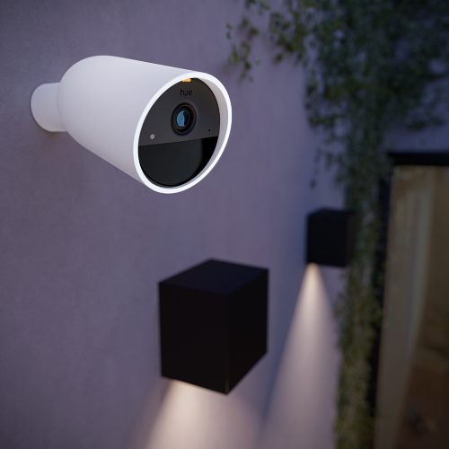 White outdoor security camera mounted on wall, lens facing forward, illuminated ring around edge, near wall lights and foliage.