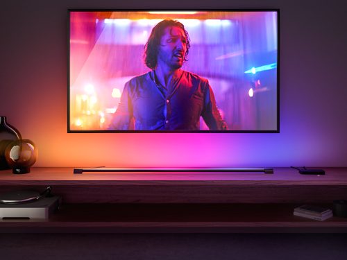 A gradient light tube placed below a wall-mounted TV casts multicolored light onto the wall, matching the on-screen content.