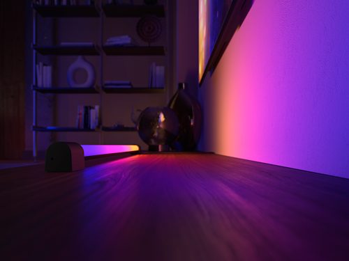 Gradient light tube placed on a wooden surface, casting multicolored illumination onto a nearby wall and tabletop objects.
